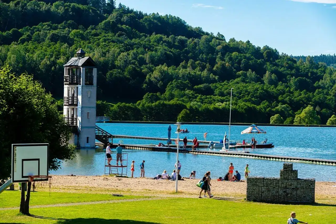 Słoneczny dzień na kąpielisku Stara Morawa. Ludzie odpoczywają na plaży, pływają i korzystają z uroków jeziora. W tle zielone wzgórza.