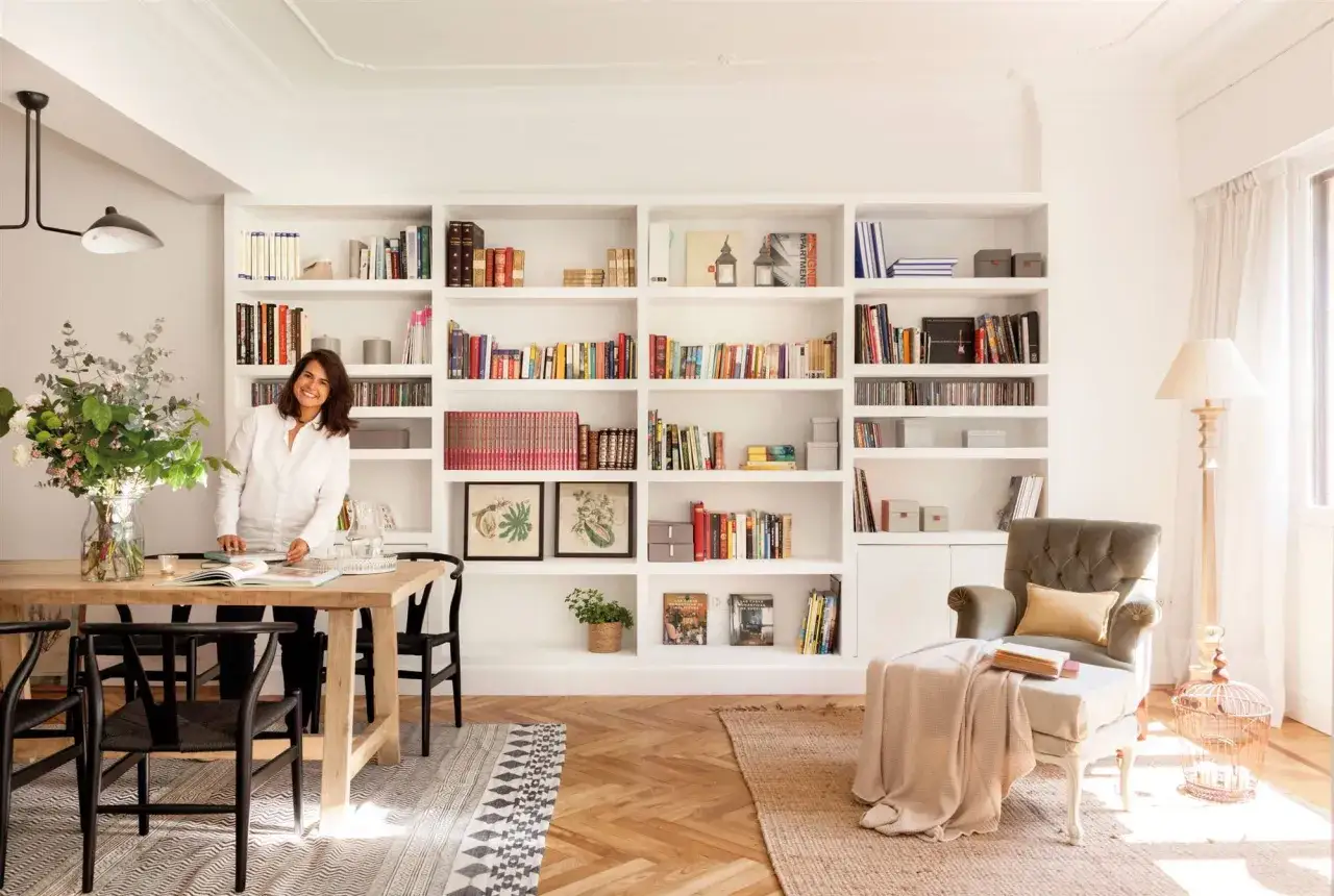 Profesional que se dedica a embellecer ambientes, sonriendo junto a una mesa de comedor y una estanter&iacute;a llena de libros.