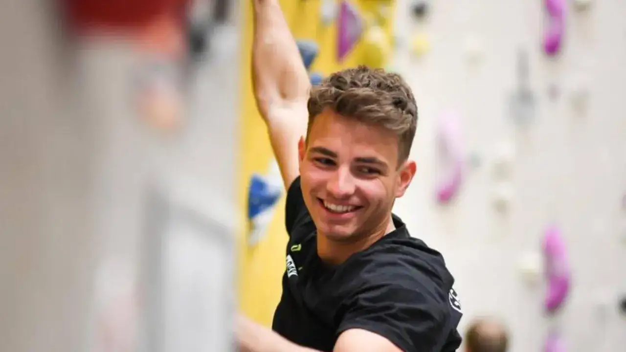 Yannick Flohé lacht, während er an einer Kletterwand hochklettert. Bunte Griffe sind an der Wand befestigt.