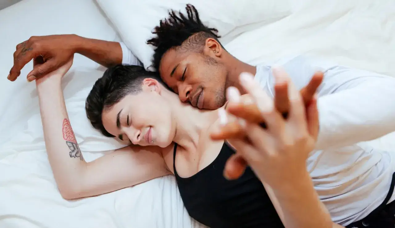 Para przytula się na ł&oacute;żku, trzymając się za ręce. Ich ciała tworzą intymne pozycje seksualne, pełne czułości i bliskości.