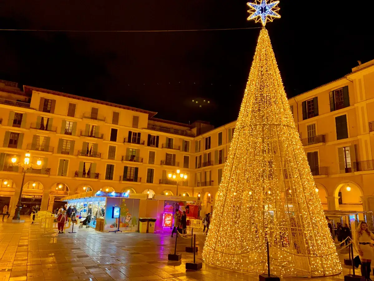 Ein riesiger, beleuchteter Weihnachtsbaum dominiert den Platz. Weihnachtliche St&auml;nde und Geb&auml;ude im Hintergrund erhellen die Nacht. Mallorca im Dezember feiert festlich.