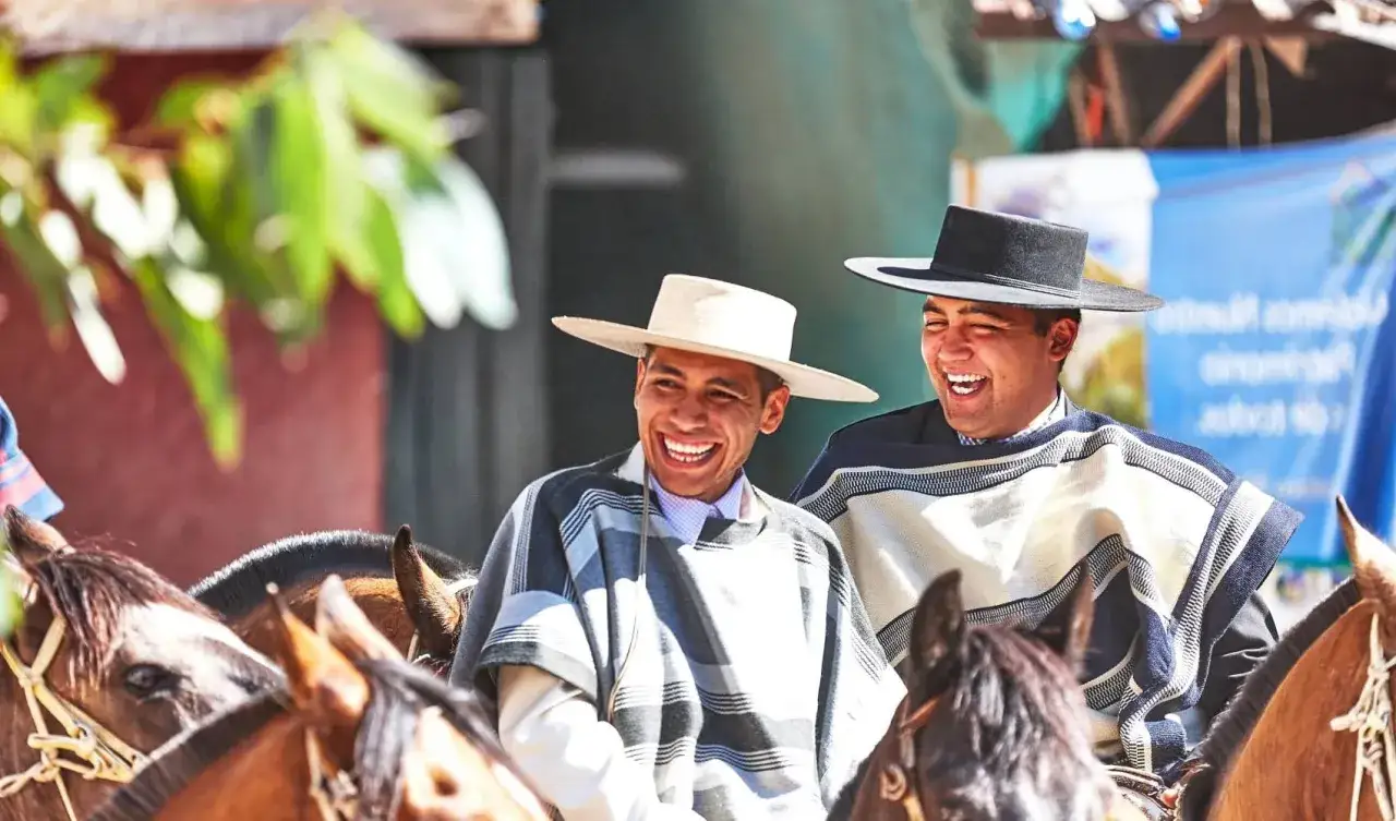 Deux huasos rient joyeusement à cheval, vêtus de ponchos traditionnels et de chapeaux.