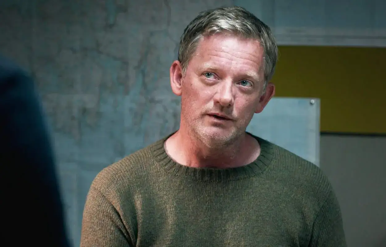 En man med grått hår och blå ögon bär en grön tröja. Han ser ut att vara mitt i ett samtal, kanske i en scen från SVT Play Shetland.