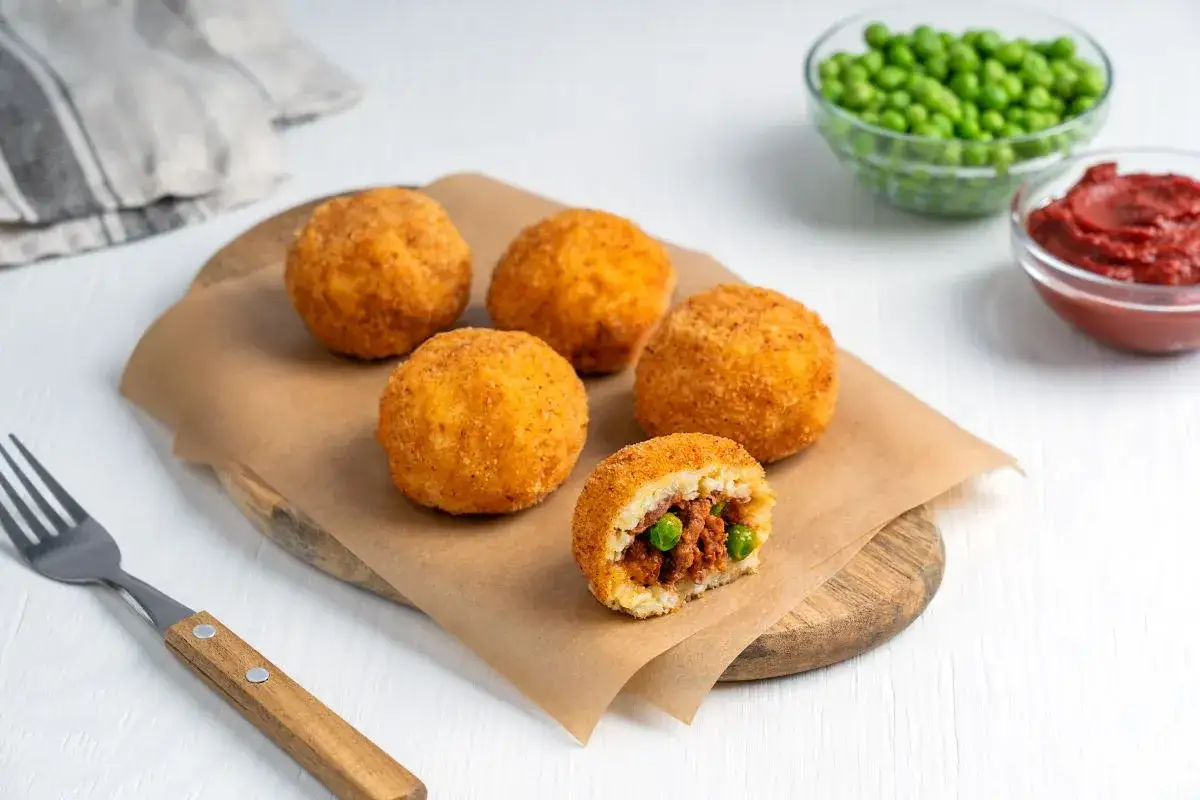 Arancini co to? Złociste kulki ryżowe z mięsem i groszkiem, jedna przekrojona, obok sos pomidorowy i groszek.