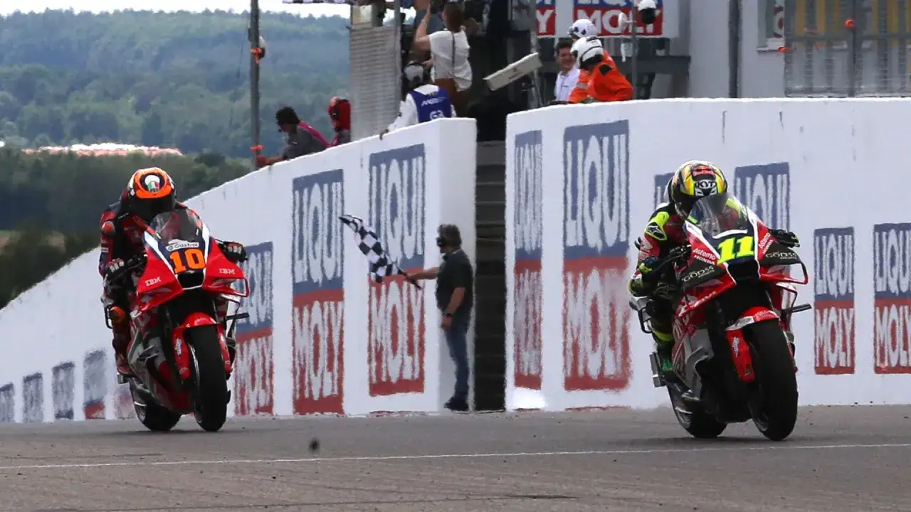Pilotos de MotoGP cruzan la meta. El número 10 lidera, mientras el 11 le sigue de cerca. Una bandera a cuadros ondea en la parrilla de salida.