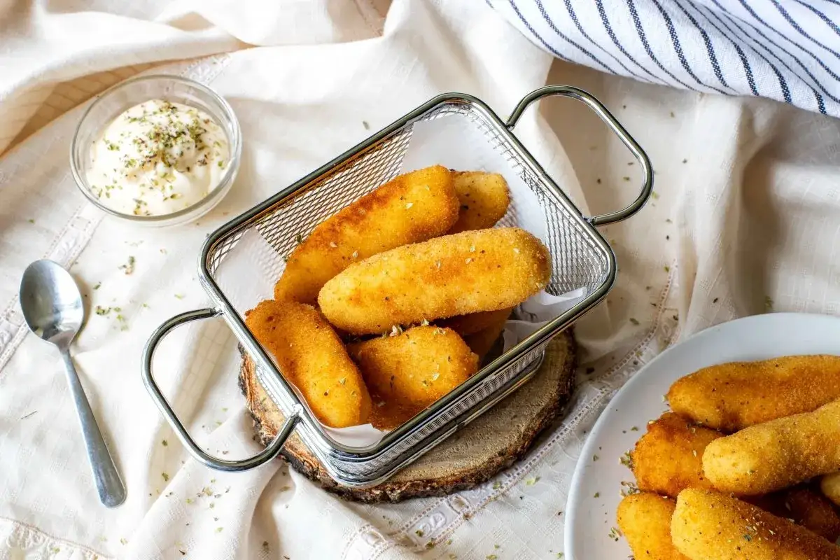 Croquetas de patata doradas y crujientes en una cesta metálica, acompañadas de una salsa cremosa.
