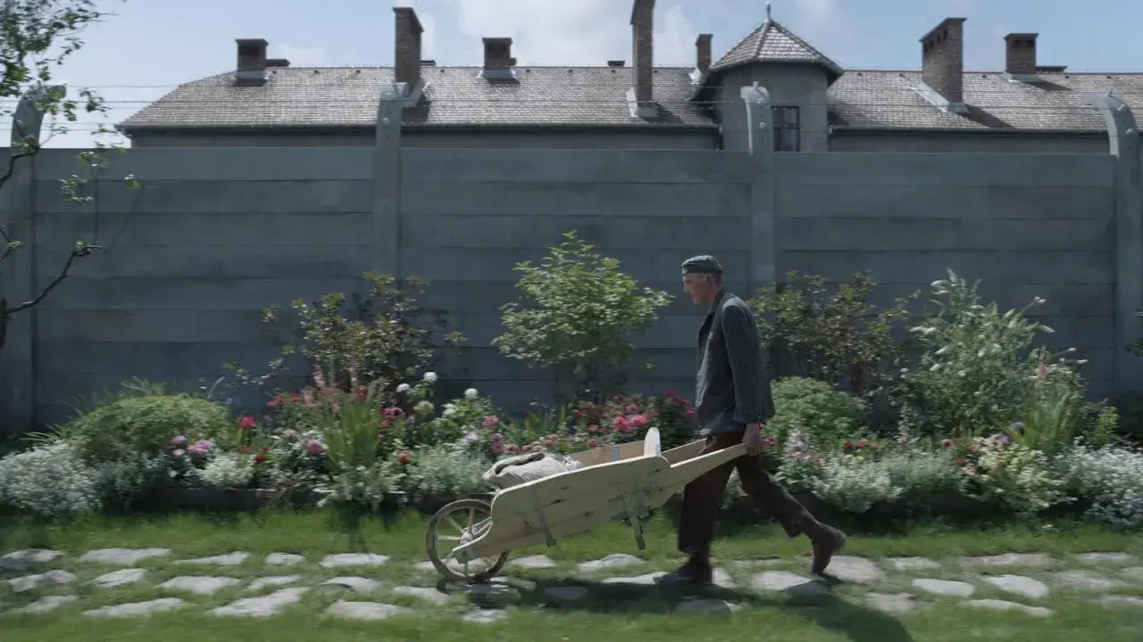 Mężczyzna pcha taczkę przez kwitnący ogród. W tle widać budynki i ogrodzenie. To scena z filmu