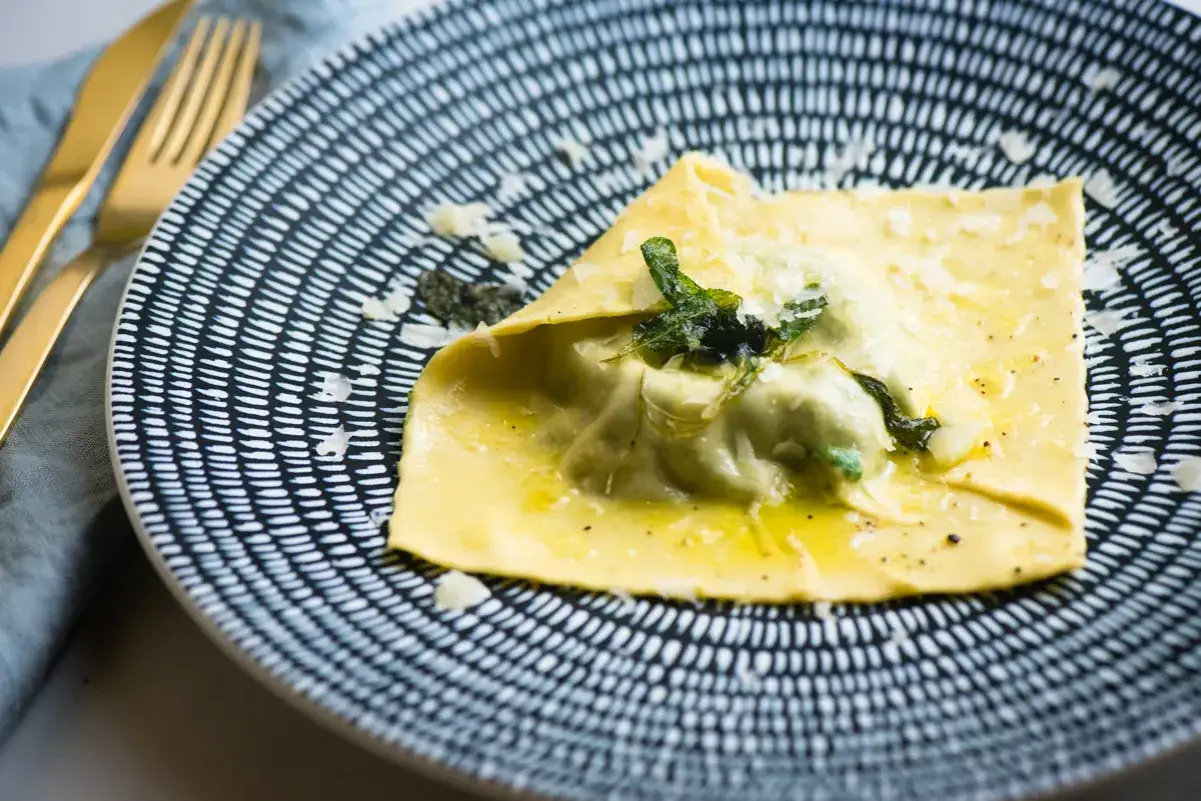 Pyszne ravioli z żółtkiem, polane masłem z szałwią i posypane parmezanem, na wzorzystym talerzu. Złote sztućce obok.