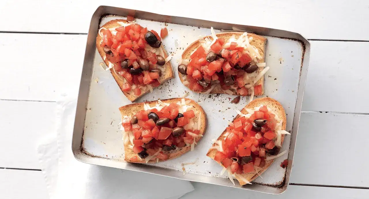 Quattro crostini pugliesi su una teglia, con pomodoro fresco, olive nere e formaggio grattugiato.