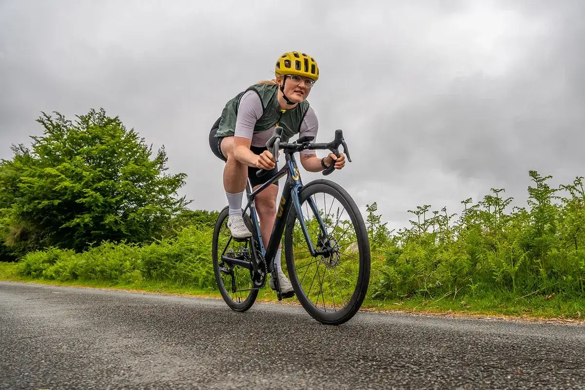Oskar Svendsen cycles intensely on a road bike, wearing a yellow helmet and a green vest, surrounded by lush greenery under a cloudy sky.