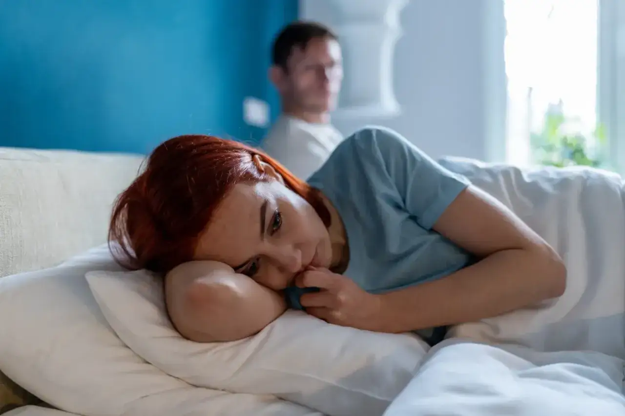 Smutna kobieta leży w ł&oacute;żku, zastanawiając się, jak zakończyć związek kiedy się kocha. Mężczyzna siedzi w tle.