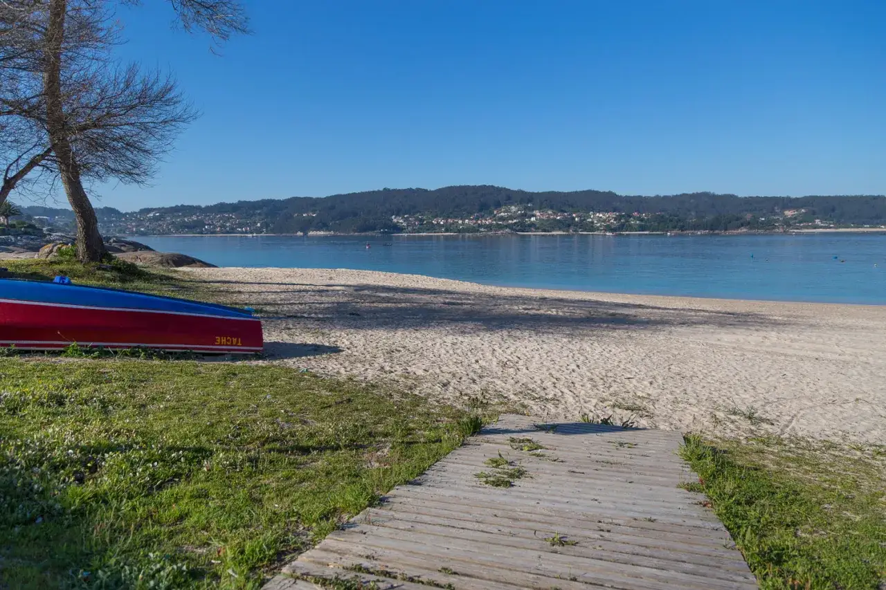 Una barca roja y azul descansa en la arena de la playa de Menduiña, con el mar tranquilo y una costa verde al fondo.