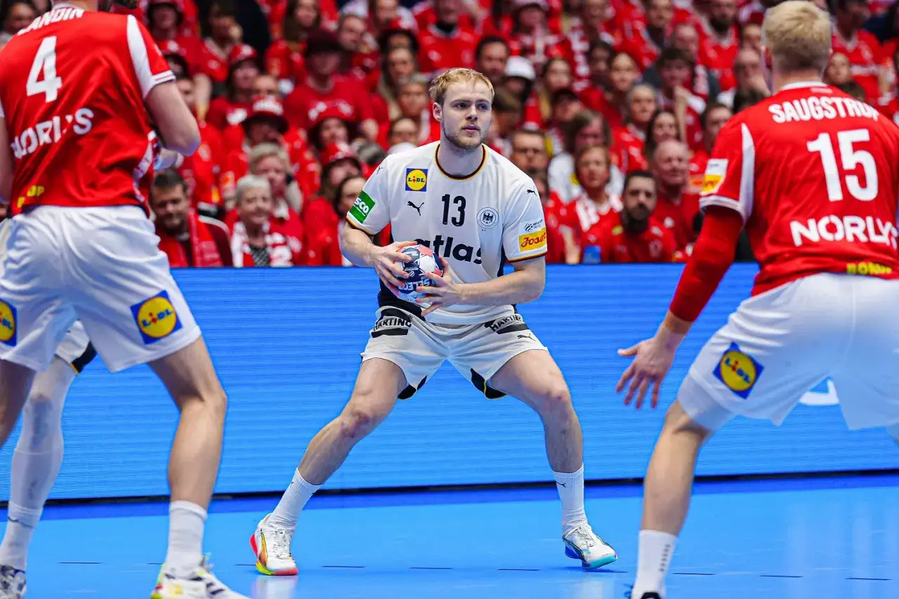 Piłkarz ręczny Niemiec z piłką, w tle kibice i zawodnicy w czerwonych strojach. Reprezentacja Danii w piłce ręcznej mężczyzn walczy o zwycięstwo.