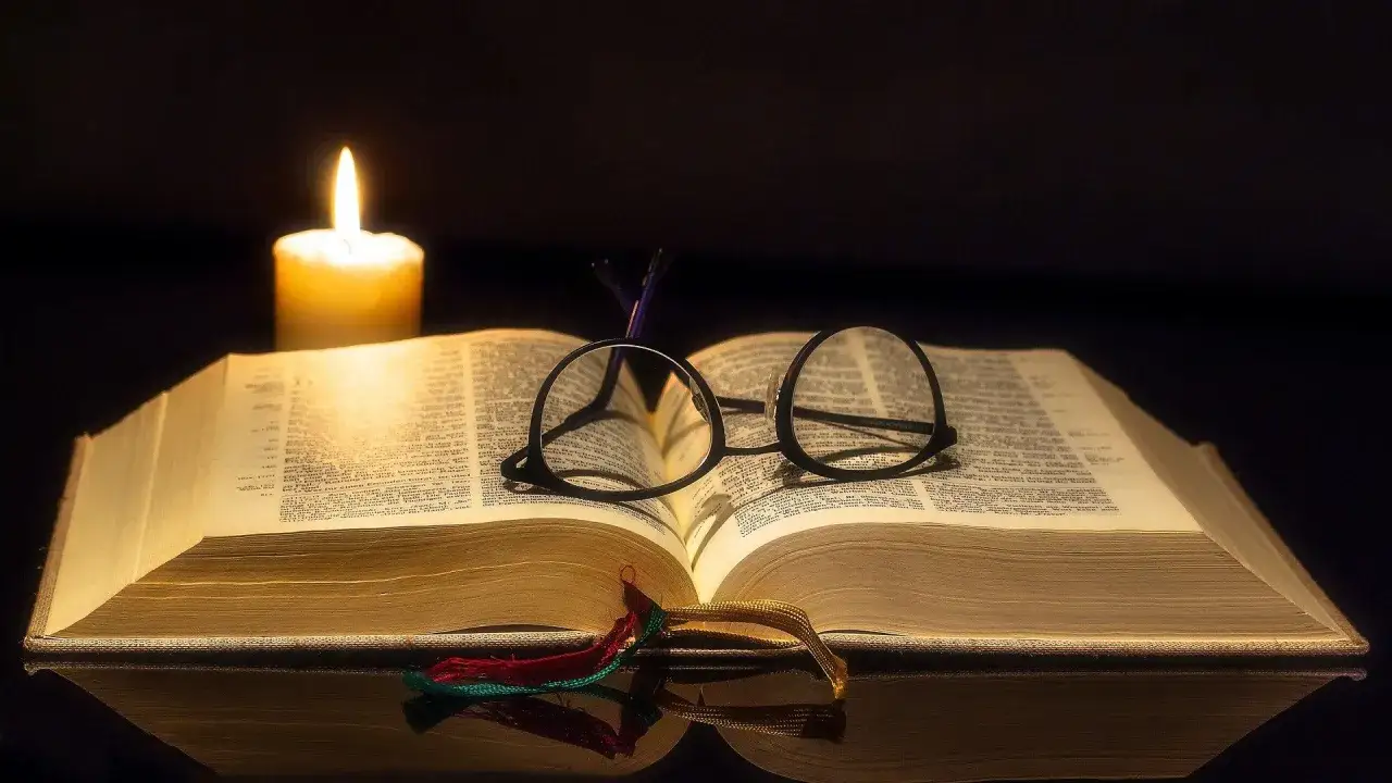 Świeca oświetla otwartą księgę, na której leżą okulary. Widać fragmenty psalmów biblijnych.