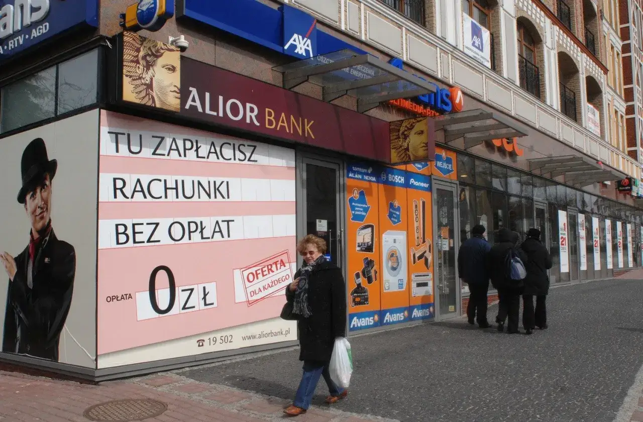Elewacja oddziału Alior Banku z reklamą "Tu zapłacisz rachunki bez opłat" i przechodzącymi ludźmi.
