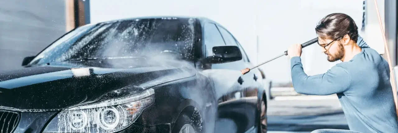 Man med glasögon tvättar en svart bil med högtryckstvätt. Vatten och skum sprutar över bilen, som om den vore en del av en kemetyl-reklam.