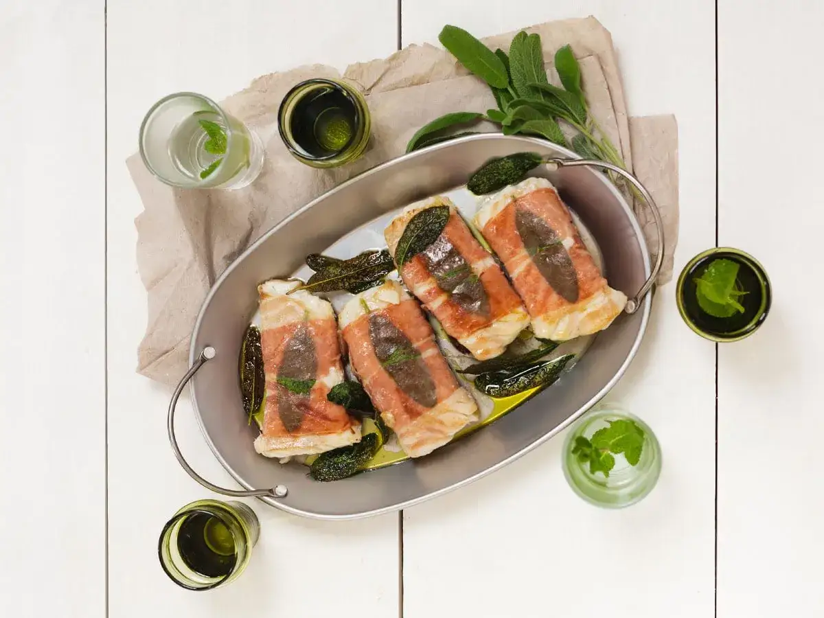 Kabeljau Saltimbocca in einer ovalen Schale, umwickelt mit Schinken und Salbei, serviert mit Kr&auml;utergetr&auml;nken.