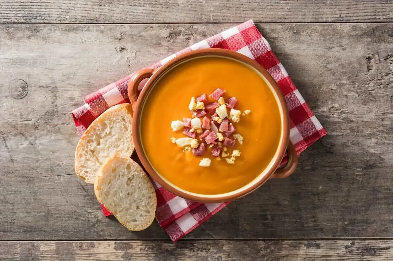 Un cuenco de salmorejo cordobés, adornado con taquitos de jamón y huevo duro, junto a rebanadas de pan rústico sobre una mesa de madera.