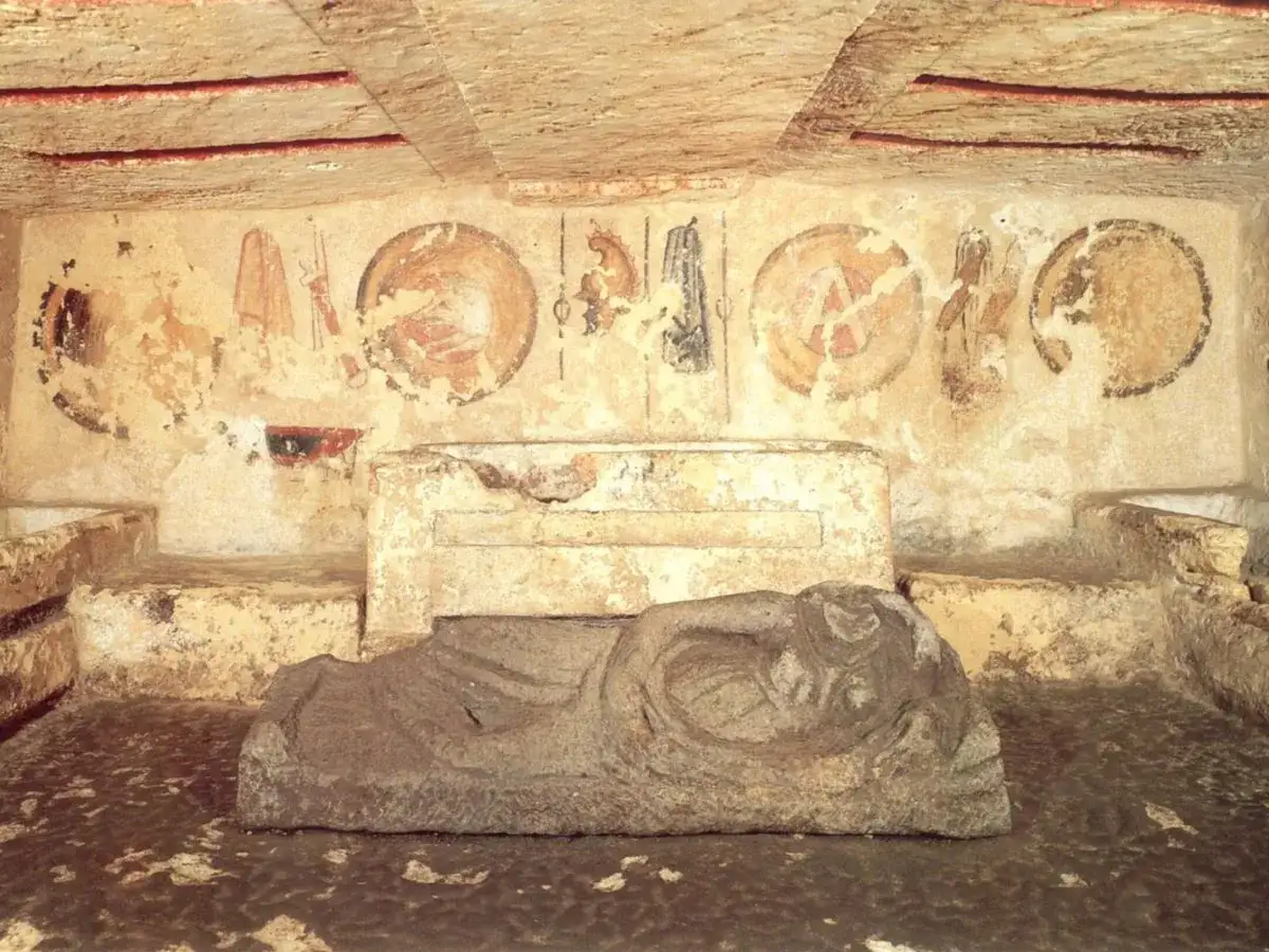Inside an Etruscan necropolis, a stone sarcophagus lies before ancient frescoes depicting shields and figures.