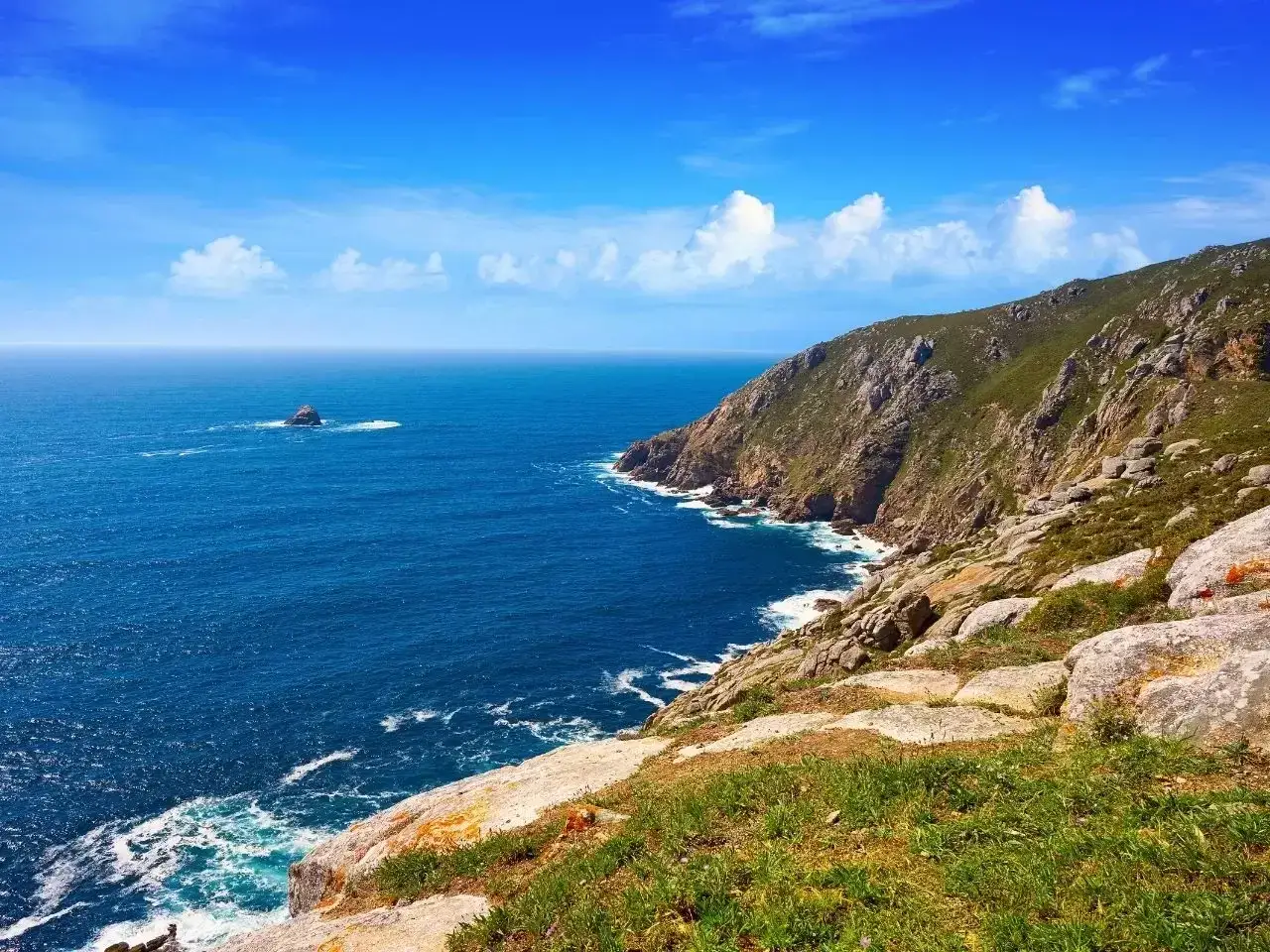 Skały i zieleń nad błękitnym oceanem, część szlaku Camino de Santiago. W oddali samotna skała.