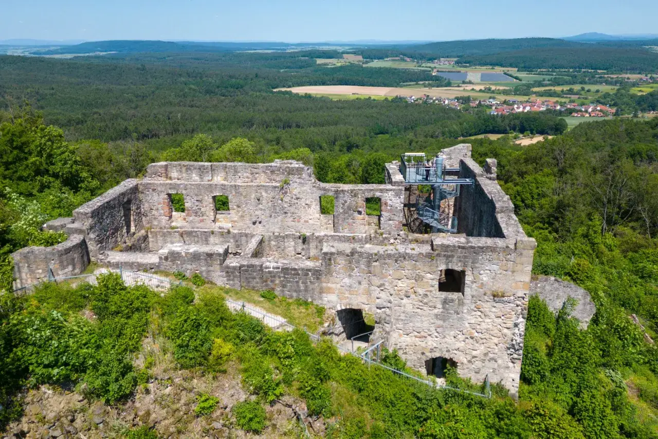 Die **Ruine Bramberg** thront auf einem bewaldeten H&uuml;gel. Steinmauern mit Fenster&ouml;ffnungen und eine Wendeltreppe sind sichtbar. Im Hintergrund erstreckt sich eine gr&uuml;ne Landschaft mit Feldern und einem Dorf.