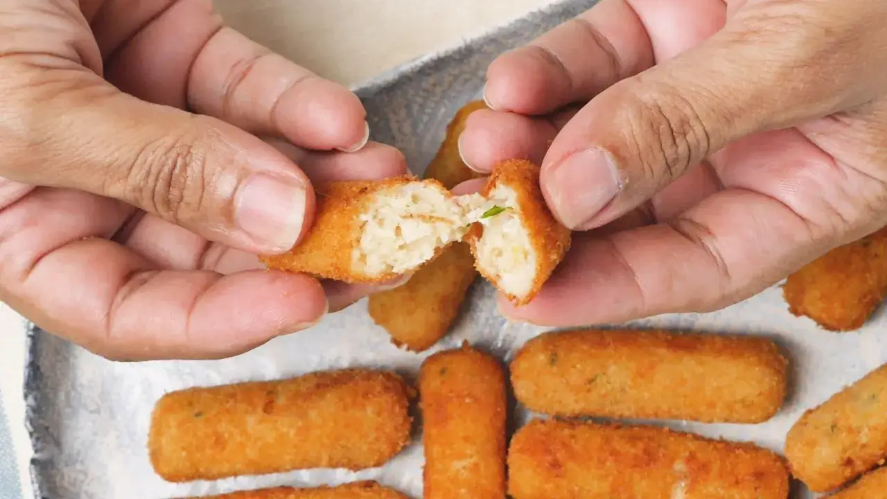Manos parten una croqueta de pollo dorada, revelando su interior cremoso. Otras croquetas esperan en una bandeja.