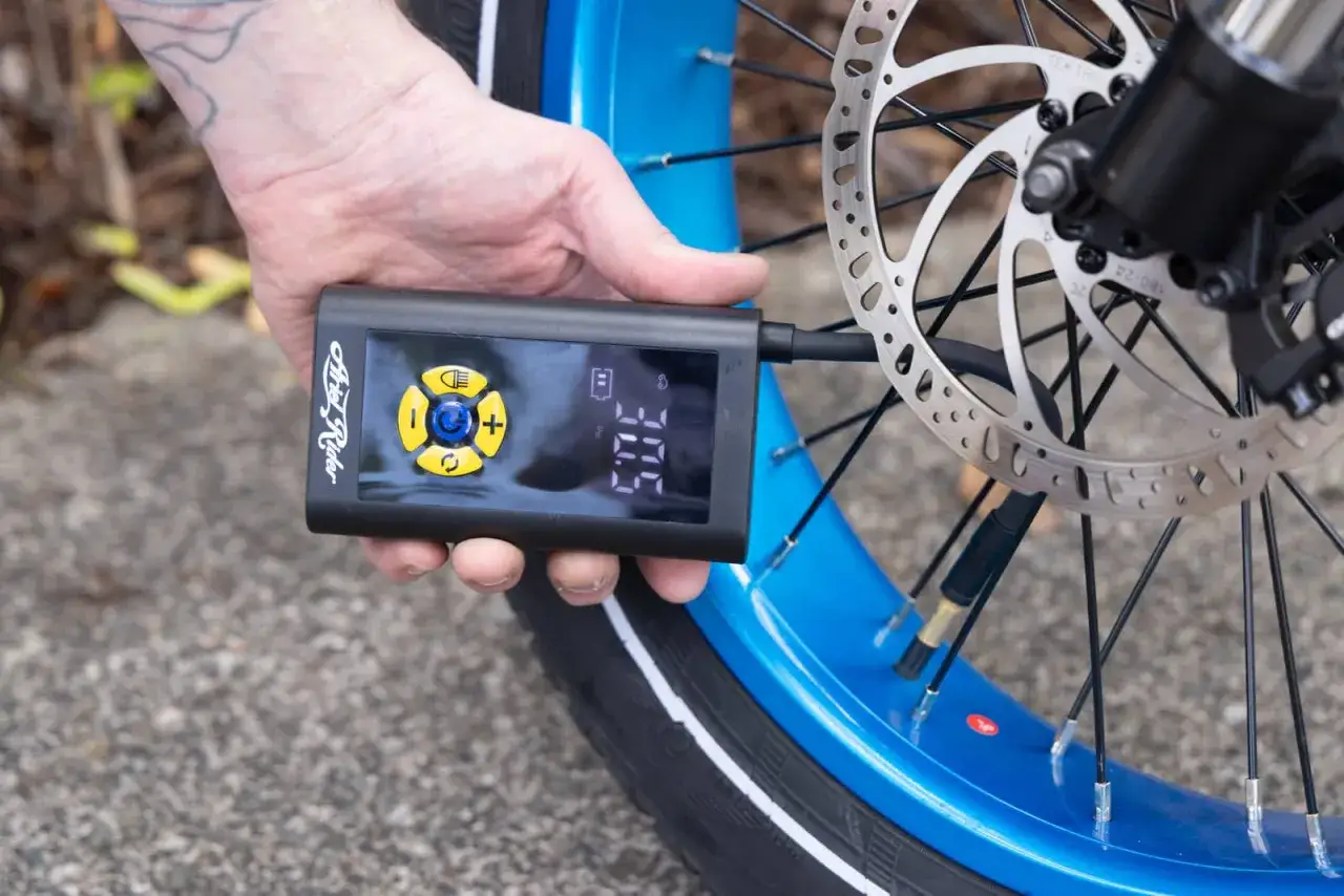 Ręka trzymająca pompkę rowerową elektryczną, która pokazuje 30.5 PSI. Urządzenie jest podłączone do wentyla niebieskiego koła roweru.