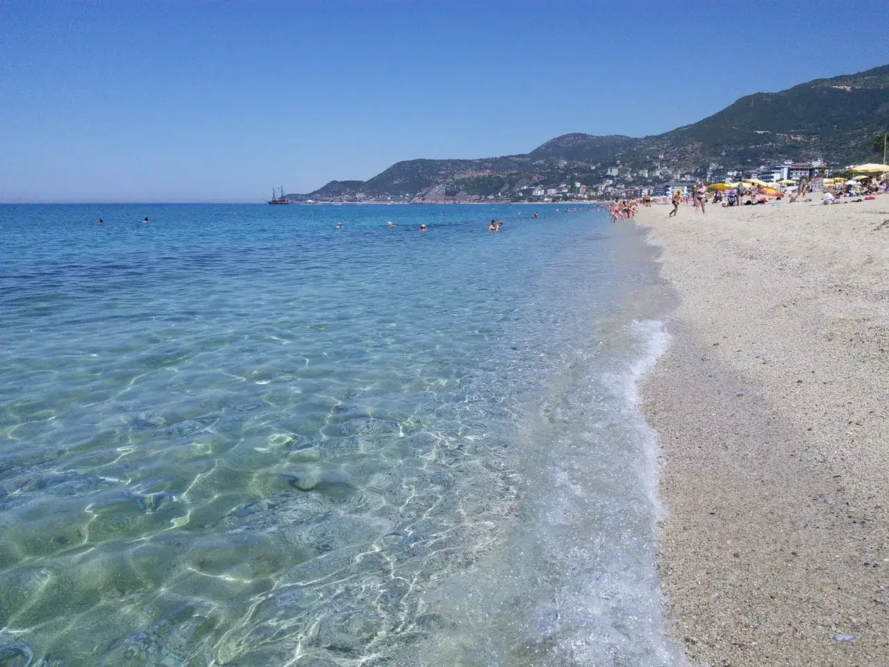 Czysta woda i złoty piasek na plaży Kleopatry. Ludzie kąpią się i opalają, a w oddali widać góry.