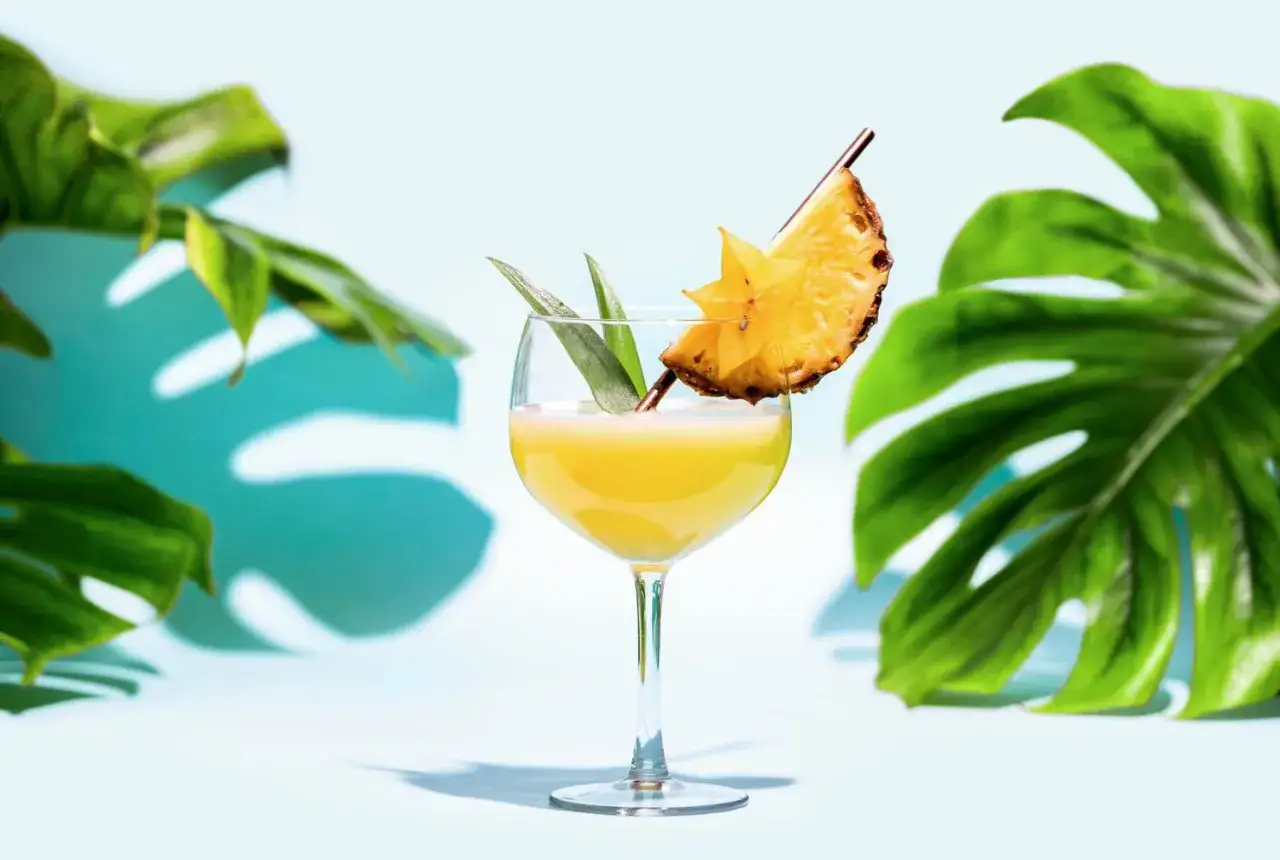 En gyllene Bahama Mama drink med ananasskiva och stjärnfrukt, omgiven av tropiska blad.