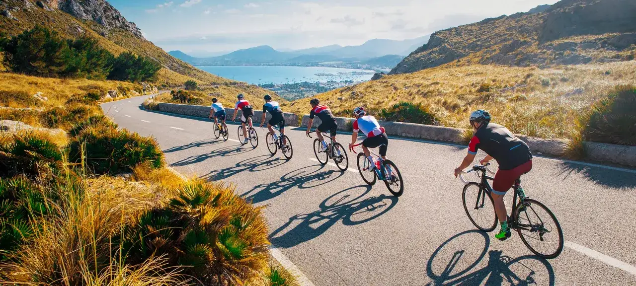 Ciclistas disfrutan de viajes en bicicleta por España, pedaleando por una carretera costera con vistas al mar y montañas.
