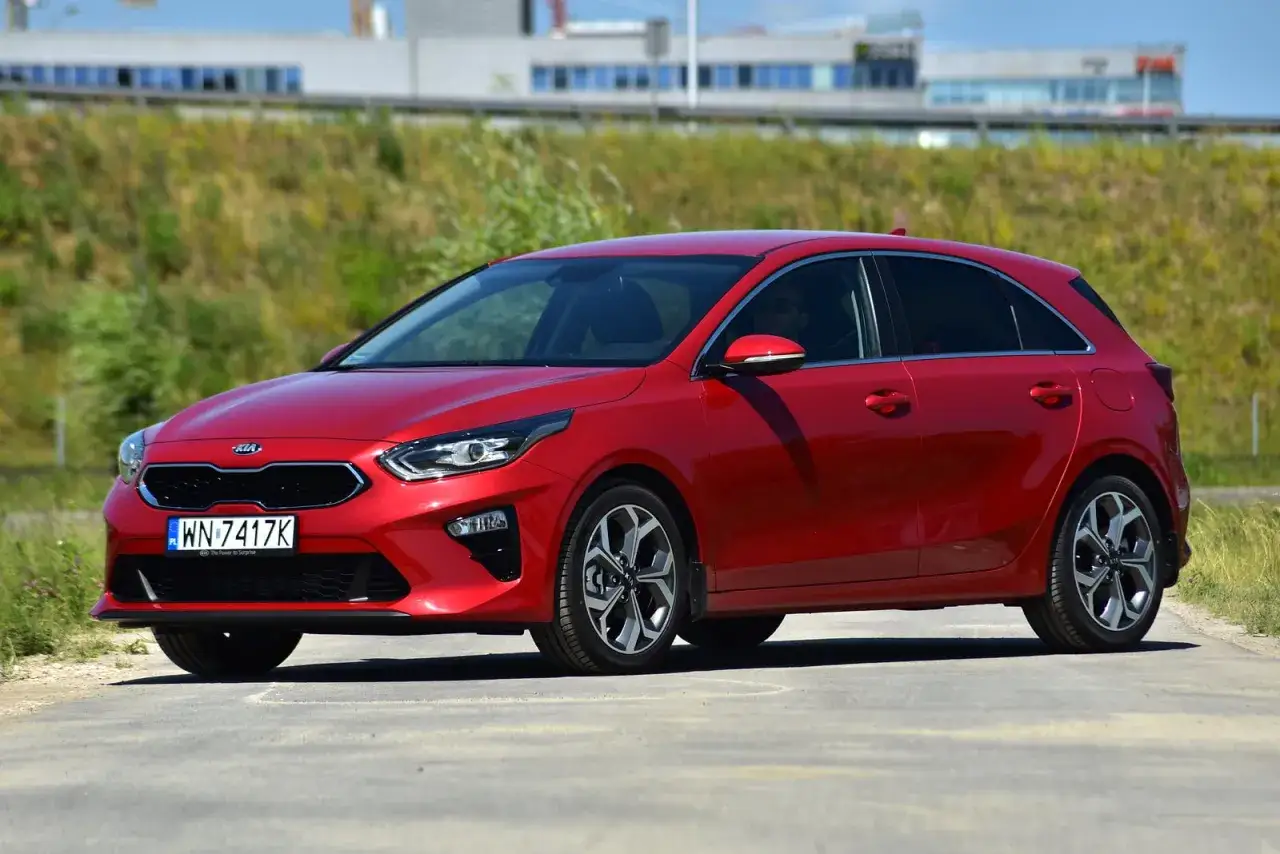 Czerwona Kia Ceed 1.6 CRDi na drodze, z zielonym pagórkiem i budynkami w tle.