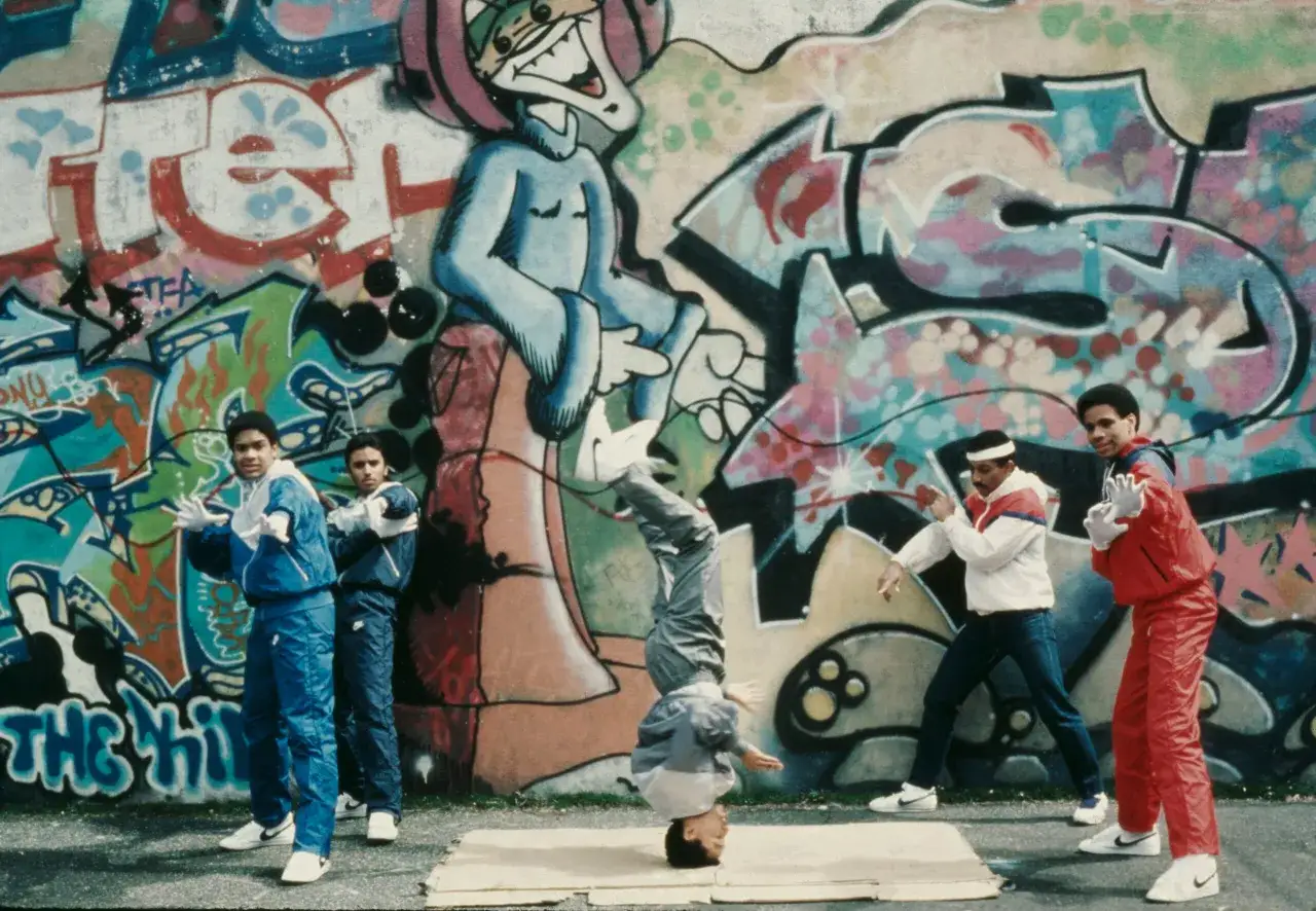 Młodzi ludzie w dresach tańczą breakdance na tle kolorowego graffiti. To esencja kultury hip hop a rap.