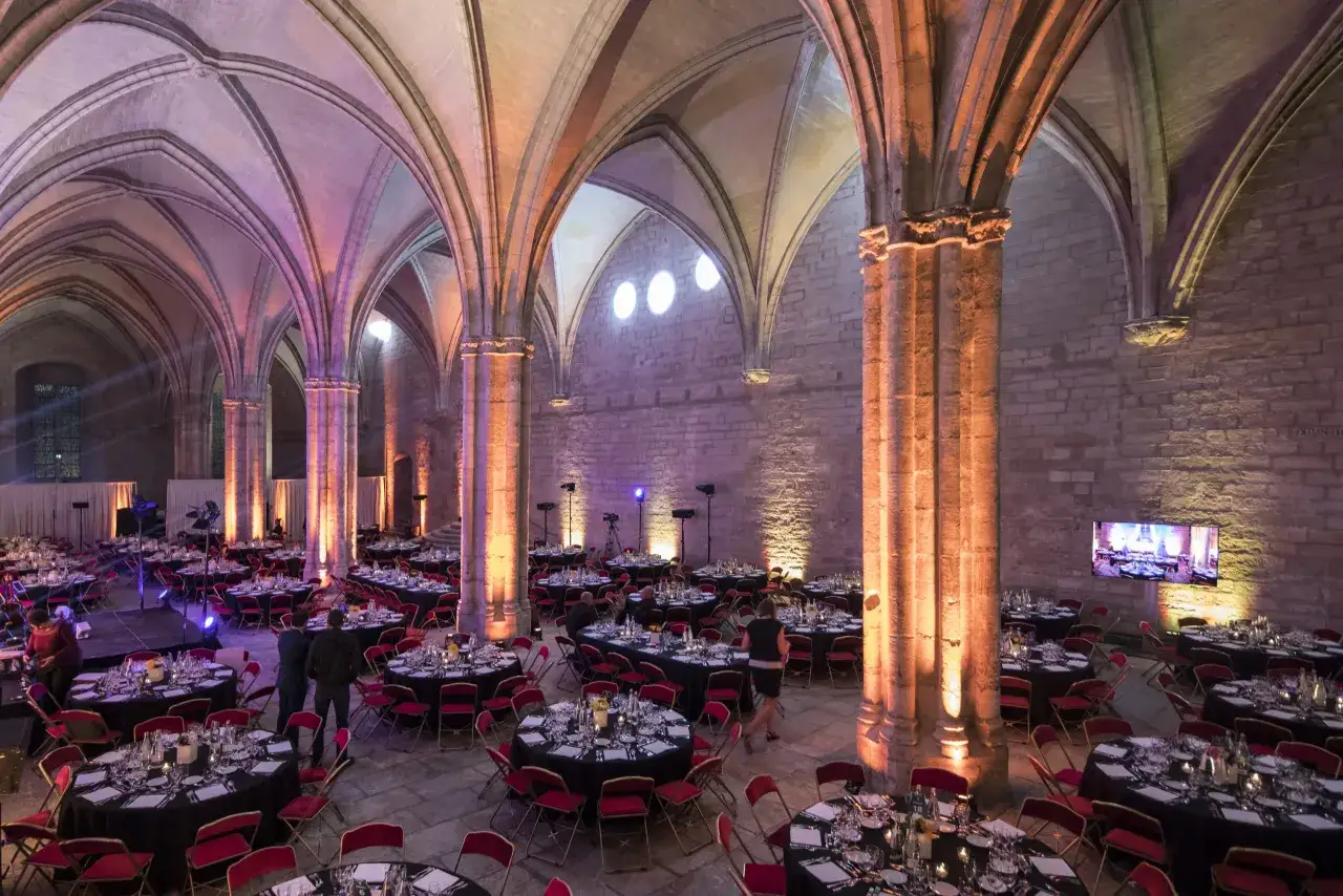 Intérieur du Palais des Papes à Avignon, une salle voûtée ornée de lumières colorées, prête pour un banquet avec des tables rondes dressées.