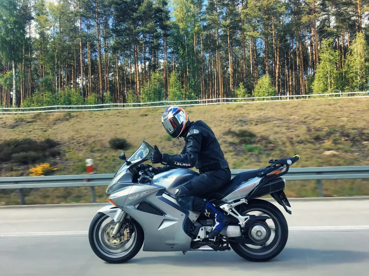 Motocyklista w kasku jedzie na srebrnym Honda VFR 800 wzdłuż leśnej drogi.