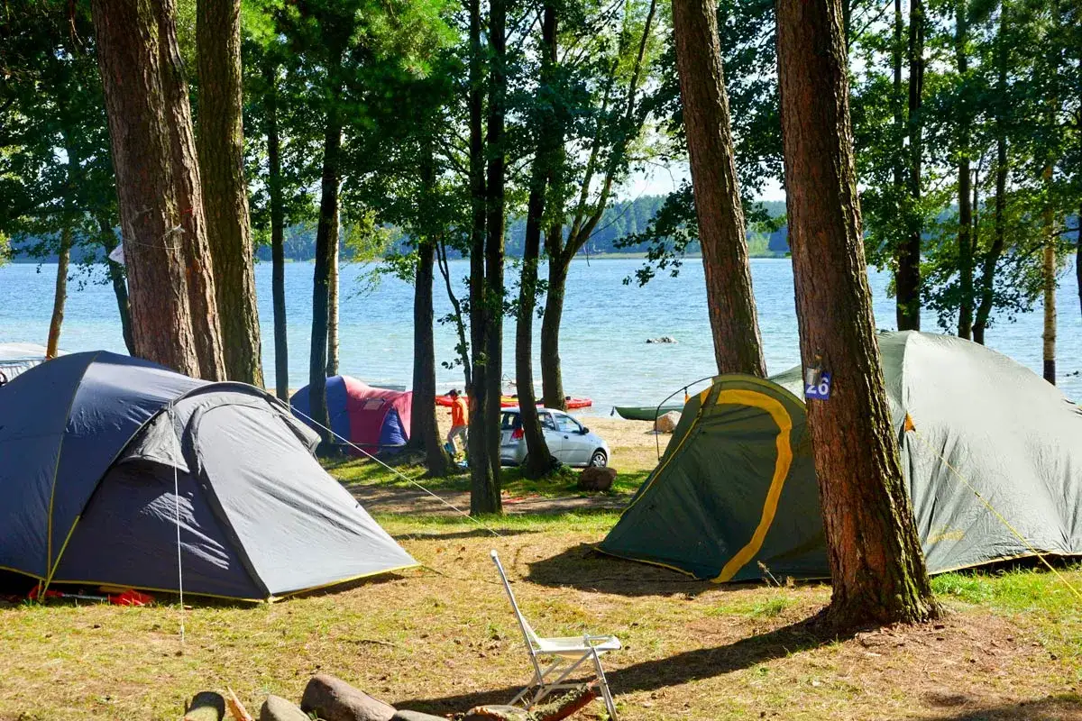 Pola namiotowe Mazury: namioty nad jeziorem, drzewa, plaża i woda.