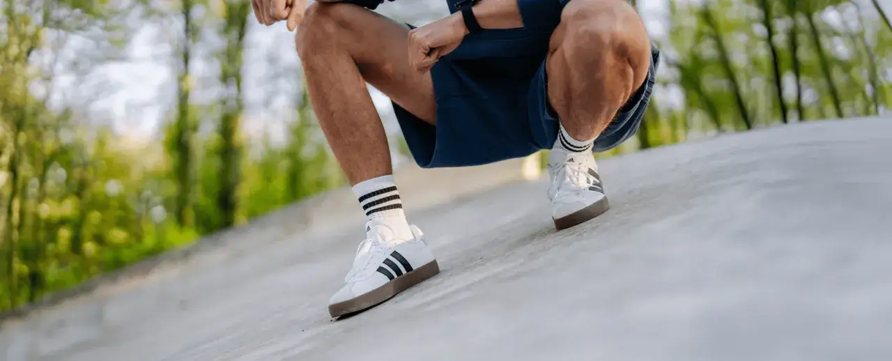 Mężczyzna w białych butach sportowych marki adidas z trzema paskami i granatowych spodenkach kuca na betonowej nawierzchni, otoczony zielenią.