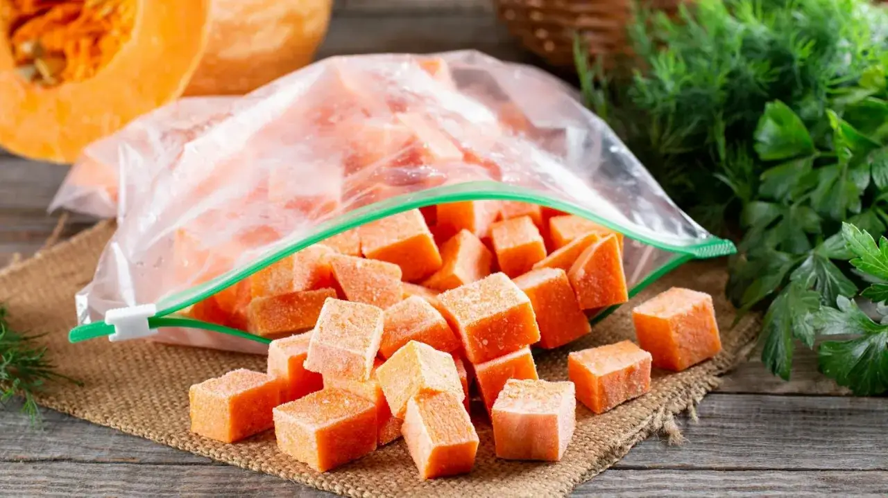 Gefrorene Würfel von Hokkaido Kürbis liegen neben einem halb geöffneten Gefrierbeutel. Frische Kräuter im Hintergrund.