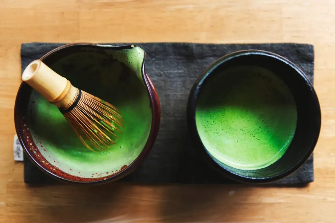 Przygotowanie idealnego matcha: bambusowa miotełka w miseczce z zielonym proszkiem i wodą, obok gotowy napój w czarnej filiżance.