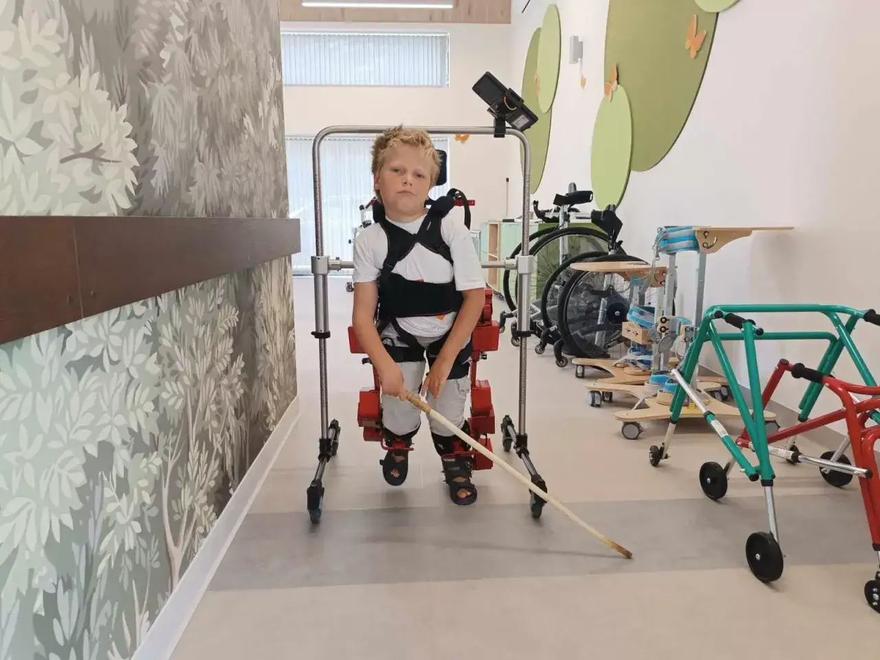 Chłopiec z rozszczepem kręgosłupa ćwiczy w specjalistycznym sprzęcie rehabilitacyjnym, wspierając się laską.