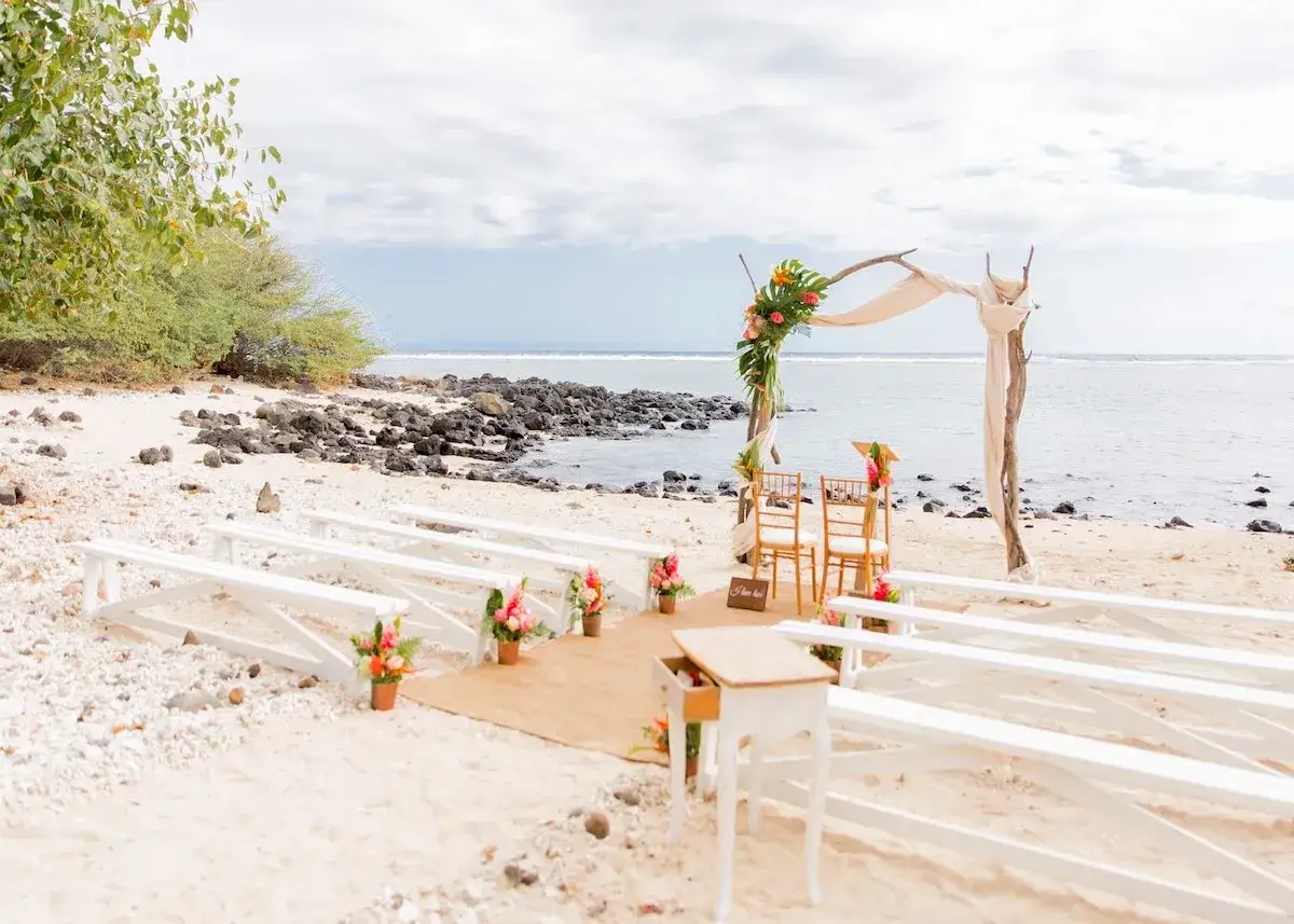 All&eacute;es de bancs blancs sur une plage de sable, d&eacute;cor&eacute;es de fleurs tropicales, pour un mariage th&egrave;me marin. L'arche de c&eacute;r&eacute;monie est orn&eacute;e de verdure et de fleurs color&eacute;es, face &agrave; l'oc&eacute;an.