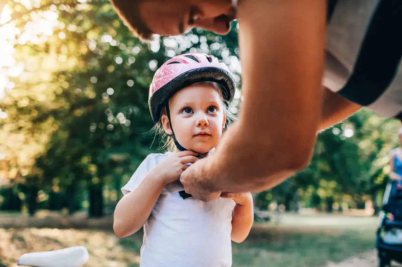 Tata zapina r&oacute;żowy kask rowerowy na głowie c&oacute;reczki. Kask w rozmiarze 52-56 cm idealnie pasuje na dziecko w wieku przedszkolnym.