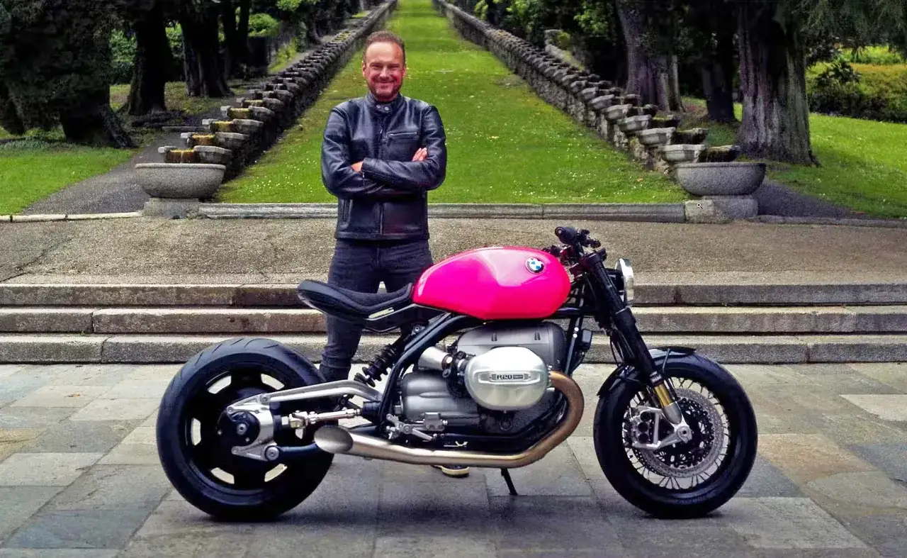 Markus Flasch lächelt neben seinem knallpinken BMW R nineT. Die Treppen und Bäume im Hintergrund bilden eine beeindruckende Kulisse.