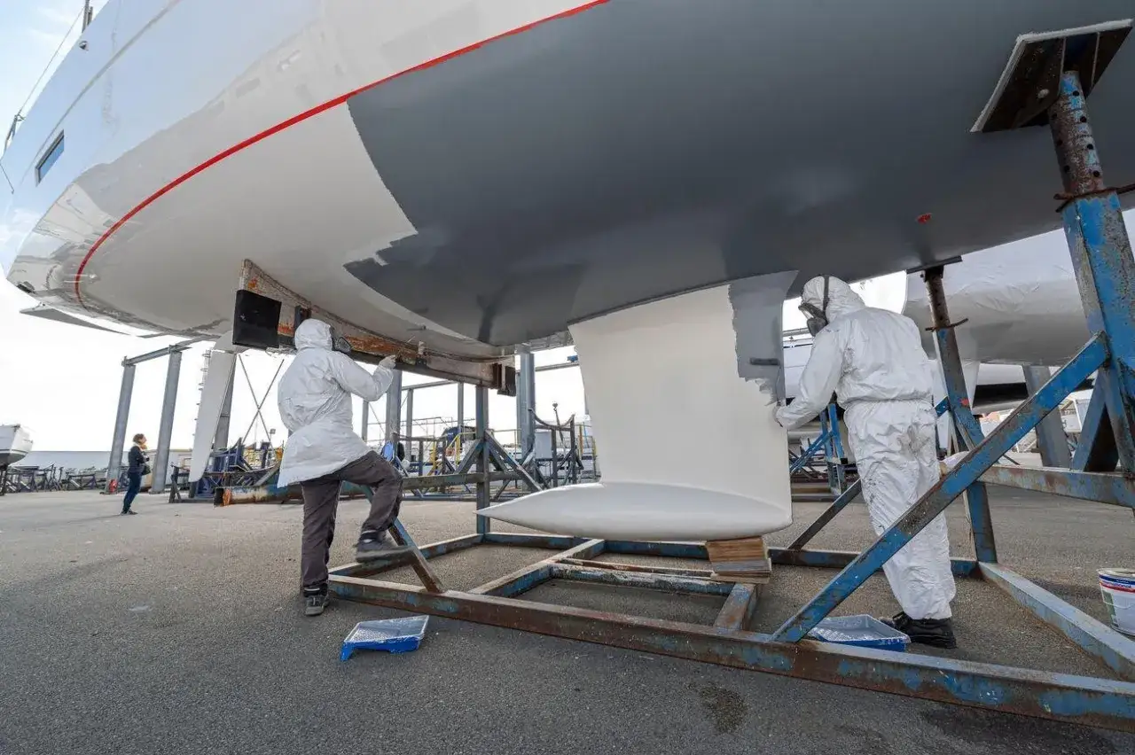 Zwei Arbeiter erneuern das Antifouling eines Bootes. Sie tragen Schutzanzüge und malen den Rumpf mit grauer Farbe.