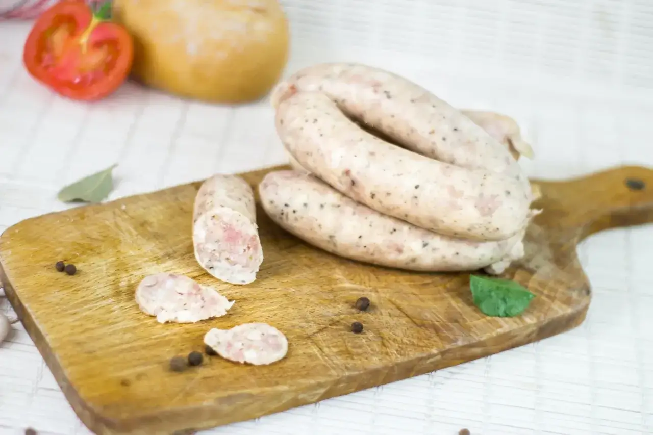 Biała kiełbasa na desce, obok pomidor, chleb i przyprawy. Wyobraź sobie moczenie jelit w occie dla tej pysznej kiełbasy.