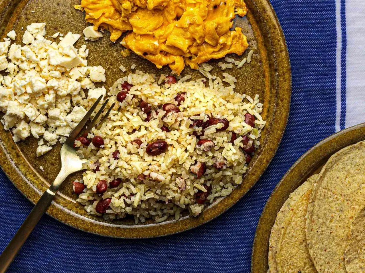 En tallrik med gallo pinto, äggröra och ost. En gaffel ligger i riset. Tortillas syns på en annan tallrik.