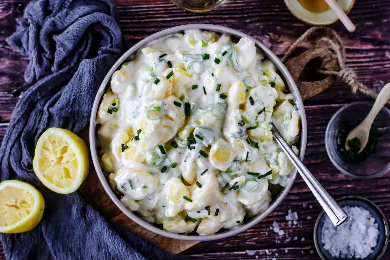 Cremiger Kartoffelsalat mit Joghurt, garniert mit Schnittlauch. Daneben liegen Zitronenh&auml;lften und ein L&ouml;ffel mit Kr&auml;utern.