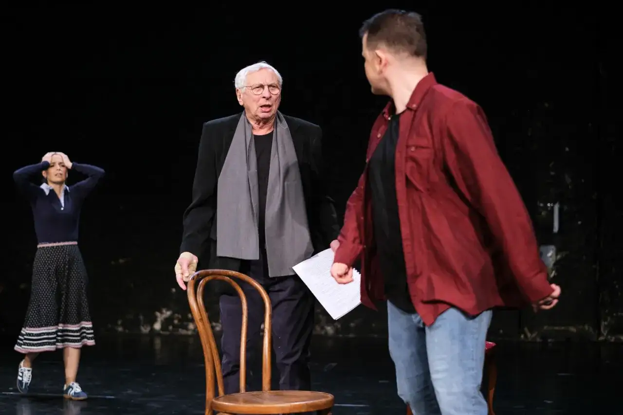 Aktor teatralny w okularach i szarym szalu rozmawia z młodszym kolegą. W tle kobieta trzyma się za głowę.