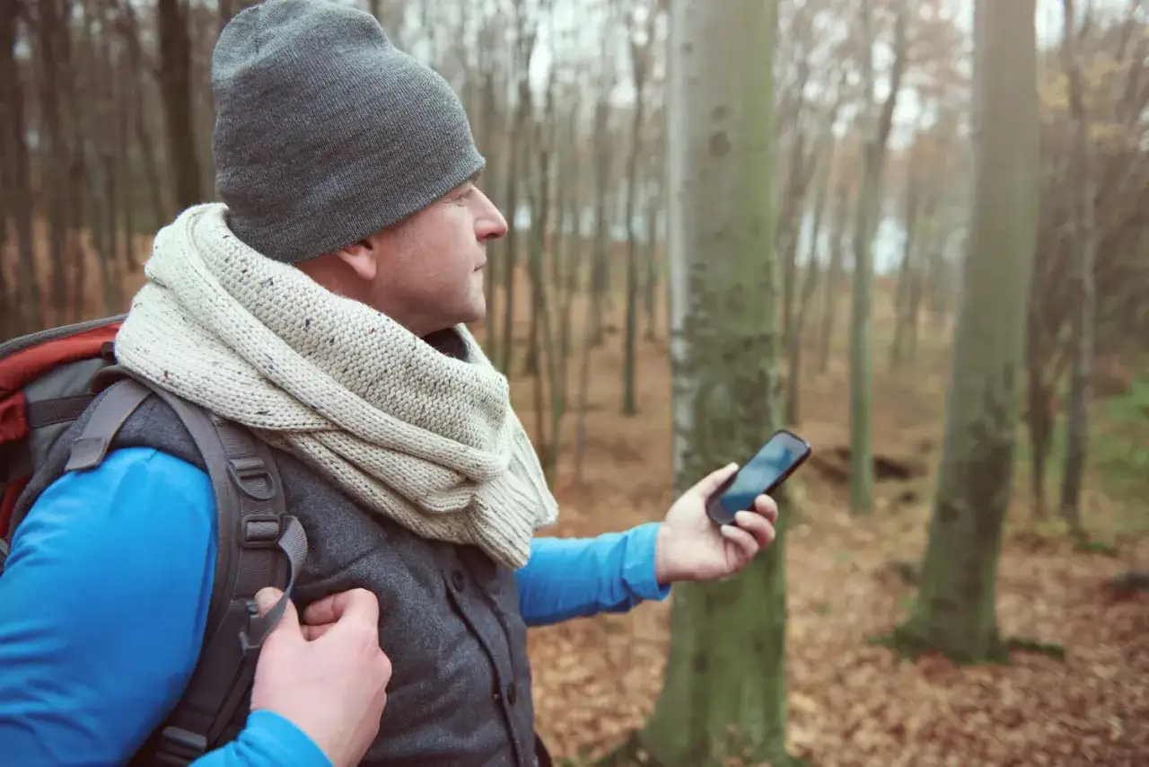 Mężczyzna w czapce i szaliku trzyma telefon, sprawdzając drogę w lesie. Wygląda jakby korzystał z nawigacji, niczym z cyfrowego kompasu.
