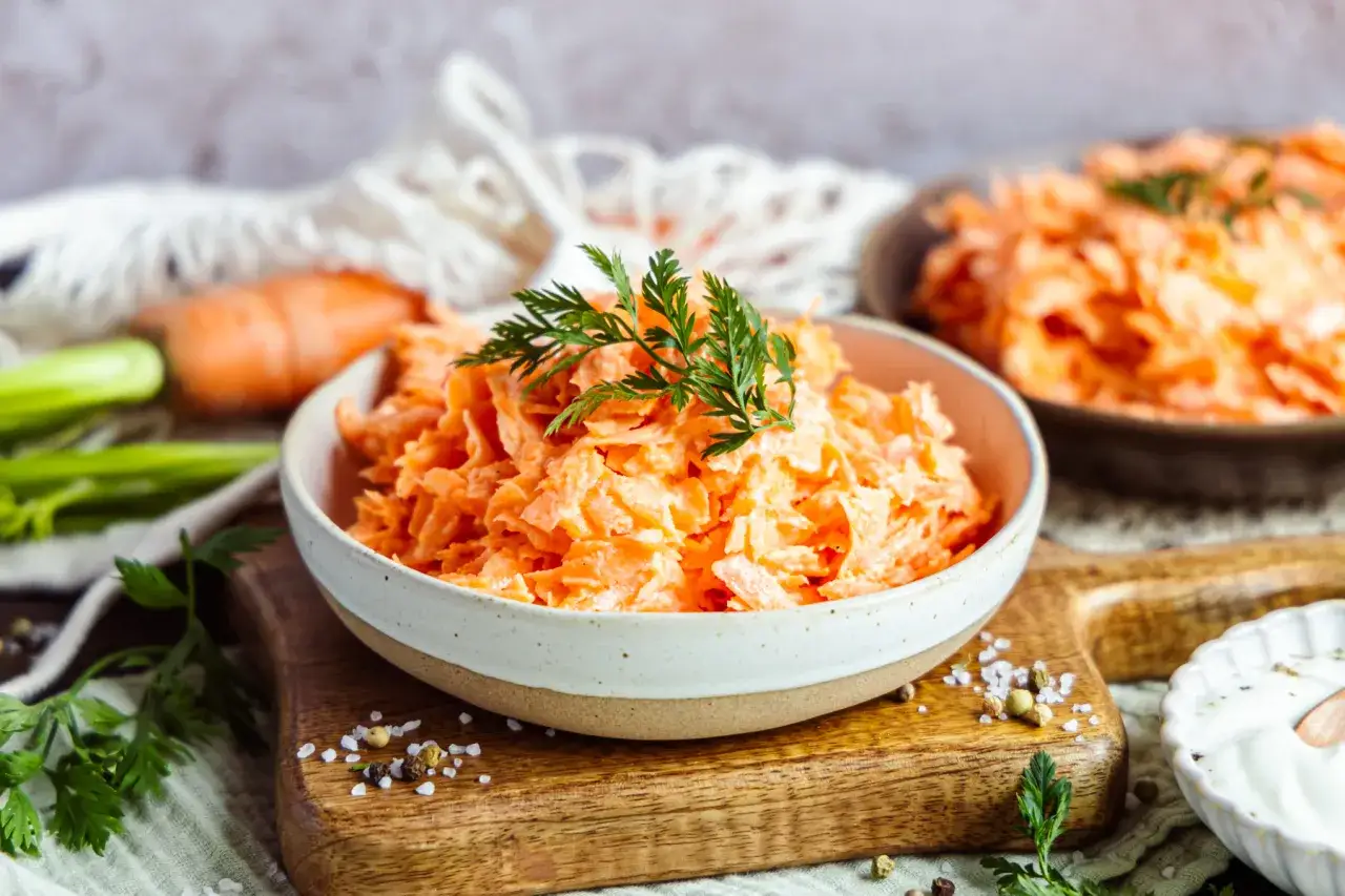 Ein cremiger Karottensalat, garniert mit Dill, perfekt für ein schnelles karottensalat rezept. Frische Karotten und Sellerie im Hintergrund.