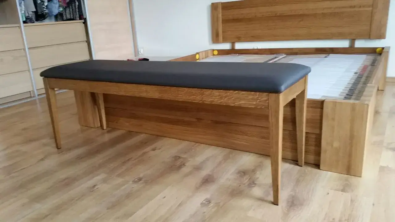 Drewniana otomana z ciemnoszarą tapicerką stoi przed łóżkiem z drewnianym zagłówkiem.